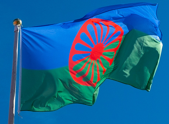 den romska flaggan mot blå himmel. Den nedre delen av flaggan är grön, den övre blå pch det är ett rött vagnshjul med många ekrar i mitten av flaggan som täcker både det gröna och det blå fältet. 