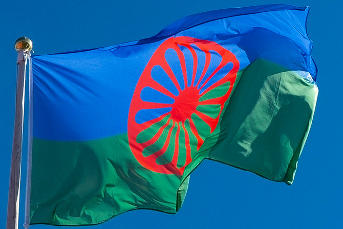 den romska flaggan mot blå himmel. Den nedre delen av flaggan är grön, den övre blå pch det är ett rött vagnshjul med många ekrar i mitten av flaggan som täcker både det gröna och det blå fältet. 