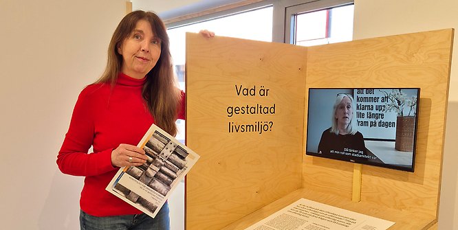 Kvinna i röd tröja som står bredvid en monter och håller en broschyr i handen.